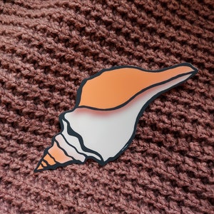 Könnte beinhalten: Ein weißer und orangefarbener Muschelaufkleber mit schwarzem Umriss. Der Aufkleber befindet sich auf einem braunen Strickstoff.