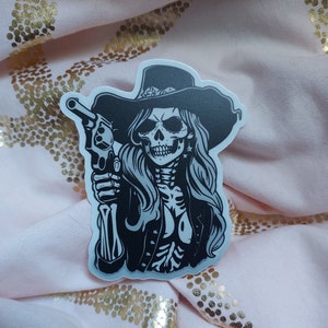 May include: Black and white sticker of a skeleton woman wearing a cowboy hat and holding a gun.