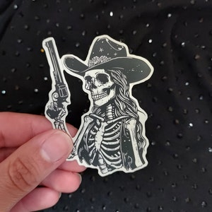 Peut inclure: Un autocollant noir et blanc d'un squelette portant un chapeau de cowboy et tenant un pistolet. Le squelette porte un gilet et a les cheveux longs.