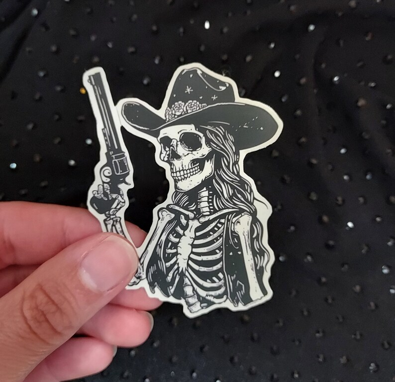 Peut inclure: Un autocollant noir et blanc d'un squelette portant un chapeau de cowboy et tenant un pistolet. Le squelette porte un gilet et a les cheveux longs.