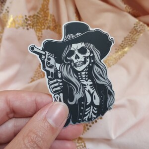May include: A black and white sticker of a skull wearing a cowboy hat and holding a gun. The skull has a long, flowing mane of hair.