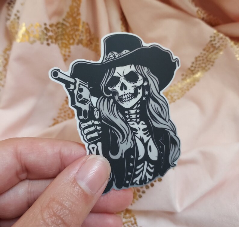 May include: A black and white sticker of a skull wearing a cowboy hat and holding a gun. The skull has a long, flowing mane of hair.