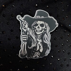 May include: Black and white sticker of a skeleton woman wearing a cowboy hat and holding a gun. The woman has long hair and is wearing a jacket.