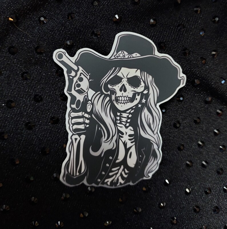 May include: Black and white sticker of a skeleton woman wearing a cowboy hat and holding a gun. The woman has long hair and is wearing a jacket.