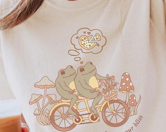 May include: Cream-colored t-shirt featuring a whimsical illustration of two green frogs riding a yellow tandem bicycle. The design includes mushrooms, flowers, and the phrase "follow your bliss." A thought bubble shows a pizza.