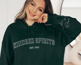 Könnte beinhalten: Ein dunkelgrüner Kapuzenpullover mit dem Schriftzug "KINDRED SPIRITS EST. 1901" in Weiß. Der linke Ärmel trägt den Schriftzug "Hope & Dream" in weißer Kursivschrift.