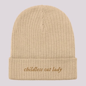 May include: Beige knitted beanie with the text "childless cat lady" embroidered in orange on the cuff.