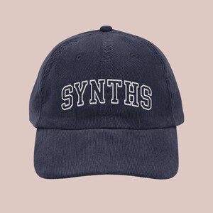 May include: A navy blue corduroy baseball cap with white embroidered text that reads "SYNTHS".