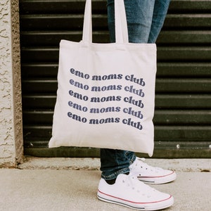May include: A cream-colored tote bag with the text "emo moms club" in a repeating, curved design in navy blue. The bag is held by a person wearing blue jeans and white sneakers with red and black trim.