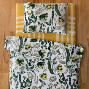 May include: A white fleece blanket with a green and yellow frog pattern. The blanket is folded in half and placed on top of a yellow and white striped fleece pad.