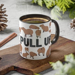 May include: A white ceramic mug with a black handle and rim. The mug has a pattern of cartoon capybaras and the text "Pull Up" in black letters.