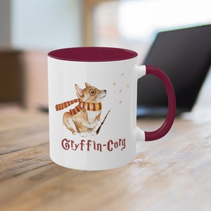 May include: White ceramic mug with a burgundy interior and handle. The mug features a watercolor illustration of a corgi wearing glasses and a scarf, holding a wand. The text "Gryffin-Corg" is printed below the corgi.