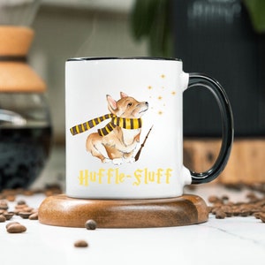 May include: White ceramic mug with a black handle and rim. The mug features a watercolor illustration of a corgi wearing a scarf and holding a wand, with the text "Huffle-Fluff" in yellow. The mug is on a wooden coaster.