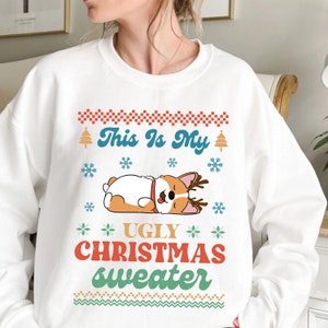 Może przedstawiać: Biały świąteczny sweter z kreskówkowym corgim w rogach i tekstem "This is my ugly Christmas sweater".