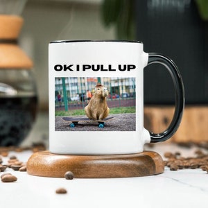 May include: White ceramic mug with a black rim and handle. The mug features a graphic of a capybara wearing a hat and riding a skateboard. The text "OK I PULL UP" is printed on the mug.