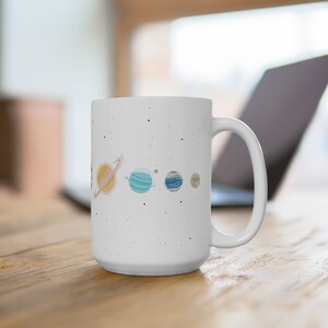 May include: White ceramic mug with a speckled finish featuring a hand-drawn illustration of the planets in our solar system. The planets are depicted in a line, with the sun on the left and the outer planets on the right. The mug has a large handle.