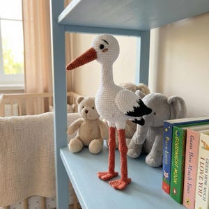 Puede incluir: Una cigüeña blanca de ganchillo hecha a mano con patas y pico naranjas y detalles de alas negras. La cigüeña está de pie sobre una estantería azul claro con otros peluches y libros. Un tema de guardería.