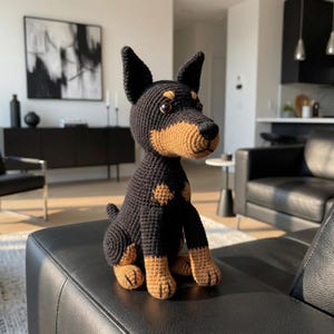 May include: A crocheted dog figurine in black and tan. The handmade plush toy features a Doberman Pinscher design with detailed stitching, including tan markings on the chest, legs, and snout. The dog is sitting on a black leather surface.