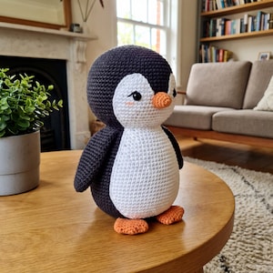 Op de afbeelding: Een handgemaakte gehaakte pinguïnpop met een zwarte kop en vleugels, een witte buik en oranje voeten en snavel. De pinguïn heeft kleine, gedetailleerde ogen en roze wangen. Hij staat op een houten tafel.