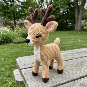 Op de afbeelding: Een handgemaakte gehaakte hertenfiguur. Het hert is voornamelijk bruin met bruine geweien en hoeven, een witte borst en roze gevoerde oren. Het heeft grote zwarte ogen en een witte snuit. Het hert staat op een houten oppervlak.