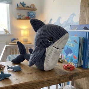 Può includere: Un giocattolo a forma di squalo fatto a mano all'uncinetto, blu e bianco. Lo squalo ha un volto sorridente e pinne dettagliate. È esposto su una mensola di legno con altre decorazioni e libri a tema oceanico.