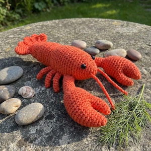Może przedstawiać: Ręcznie robiona, jasnoczerwona szydełkowa homar z czarnymi oczami, długimi czułkami i szczegółowymi szczypcami. Homar leży na kamiennej powierzchni z małymi kamykami i zielenią w tle. Unikalny, ręcznie robiony przedmiot o tematyce morskiej.