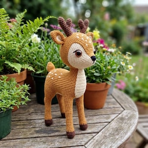 May include: A handmade crochet deer figurine with brown and cream colors. The deer has brown antlers, dark brown hooves, and white spots. It is standing on a wooden surface, with potted plants in the background.