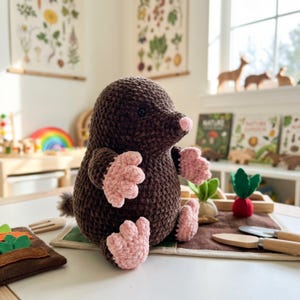 May include: A handmade brown crochet mole plush toy with pink paws and nose. The plush is sitting on a table with other wooden toys and a felt garden set. The background includes botanical prints and a window.