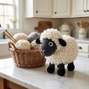 以下が含まれることがあります： ふわふわの白い体、黒い顔、耳、足、ピンクの鼻を持つ手作りのかぎ針編みの羊。その隣には、毛糸玉と編み針が入った柳のバスケットがあり、手芸プロジェクトに最適です。