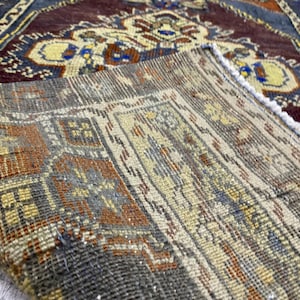 May include: A folded woven rug featuring a complex geometric pattern in shades of deep burgundy, cream, orange, and blue. The underside reveals a coarse, textured weave with visible fibers, while the top side displays a detailed floral motif.