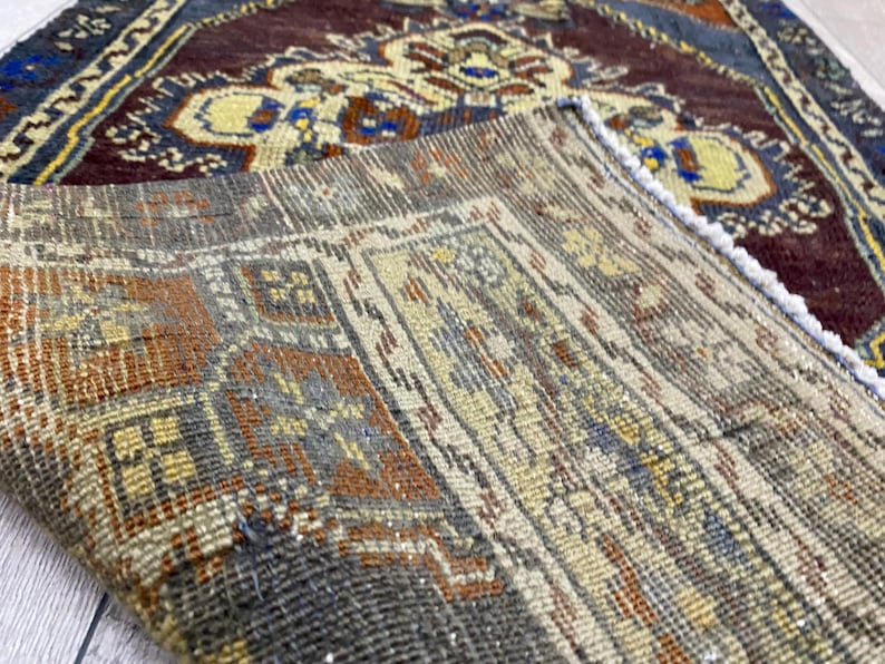May include: A folded woven rug featuring a complex geometric pattern in shades of deep burgundy, cream, orange, and blue. The underside reveals a coarse, textured weave with visible fibers, while the top side displays a detailed floral motif.