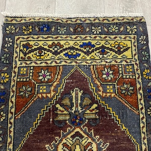May include: A rectangular woven rug featuring a central dark maroon mihrab design surrounded by a deep gray field. The border displays intricate floral motifs in yellow, blue, and green, finished with a short white fringe at the top edge.