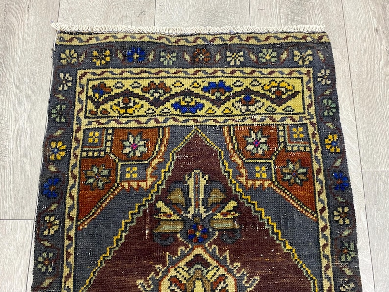 May include: A rectangular woven rug featuring a central dark maroon mihrab design surrounded by a deep gray field. The border displays intricate floral motifs in yellow, blue, and green, finished with a short white fringe at the top edge.
