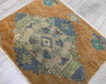 Puede incluir: Una alfombra rectangular con un diseño de medallón central en tonos beige, azul, verde y morado, sobre un fondo naranja cálido. La alfombra tiene una textura de pelo corto y flecos blancos en los bordes superior e inferior.