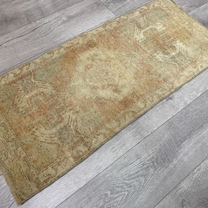 Puede incluir: Una alfombra rectangular con un patrón desgastado y descolorido en tonos beige, bronceado y naranja apagado. La alfombra presenta un diseño de medallón central con bordes geométricos intrincados y flecos cortos deshilachados en ambos extremos.
