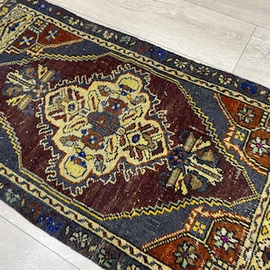 May include: A rectangular area rug featuring a deep burgundy central medallion surrounded by a dark charcoal field. The design includes intricate geometric patterns in shades of yellow, blue, and rust orange, finished with a short fringe on the ends.