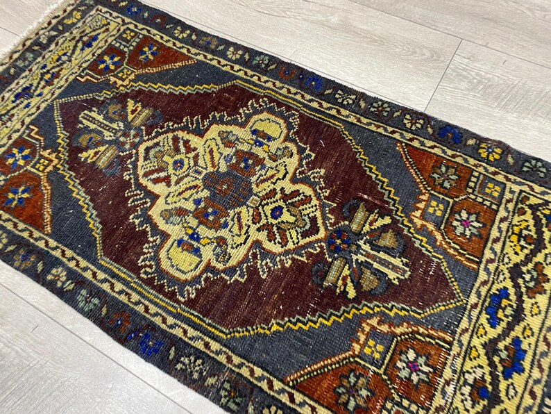 May include: A rectangular area rug featuring a deep burgundy central medallion surrounded by a dark charcoal field. The design includes intricate geometric patterns in shades of yellow, blue, and rust orange, finished with a short fringe on the ends.
