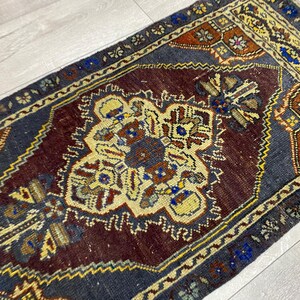 May include: A rectangular area rug featuring a deep maroon central medallion surrounded by a dark blue field. The design includes intricate geometric patterns in yellow, orange, and blue, with a decorative border and short white fringe at the ends.