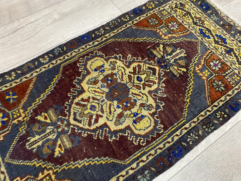 May include: A rectangular area rug featuring a deep maroon central medallion surrounded by a dark blue field. The design includes intricate geometric patterns in yellow, orange, and blue, with a decorative border and short white fringe at the ends.