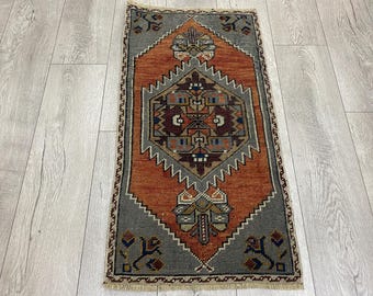 May include: A rectangular, hand-woven rug with a central diamond pattern in shades of orange, brown, and beige. The rug features a gray border with floral motifs and a fringed edge. The rug's dimensions are approximately 24 x 36 inches.