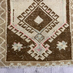 May include: A small, rectangular rug with a central diamond pattern in shades of brown and cream. The rug features geometric designs and a fringed edge. The border is a lighter tan color, and the rug has a vintage, handcrafted appearance.