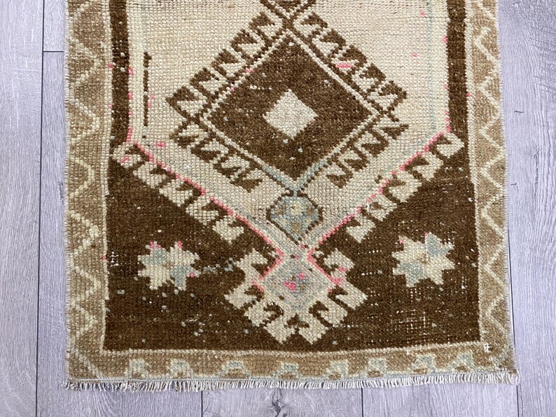 May include: A small, rectangular rug with a central diamond pattern in shades of brown and cream. The rug features geometric designs and a fringed edge. The border is a lighter tan color, and the rug has a vintage, handcrafted appearance.