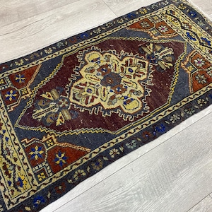 May include: A rectangular area rug featuring a central dark maroon medallion surrounded by a dark blue field. The border displays intricate geometric patterns in yellow, red, and blue. The rug has short white fringe on the ends and a woven texture.