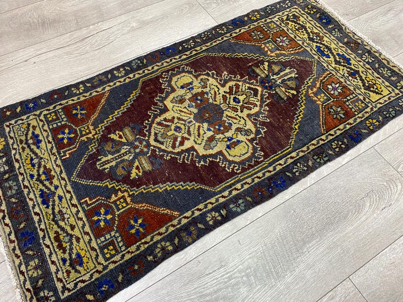 May include: A rectangular area rug featuring a central dark maroon medallion surrounded by a dark blue field. The border displays intricate geometric patterns in yellow, red, and blue. The rug has short white fringe on the ends and a woven texture.