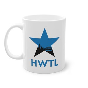 May include: White ceramic mug with a blue star design and a black cityscape silhouette. The mug features the blue letters "HWTL". The mug has a curved handle.