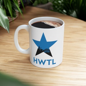 May include: White ceramic mug featuring a blue star design. Inside the star is a black silhouette of a city skyline and bridge. The letters "HWTL" are printed below the star. The mug is filled with dark coffee.
