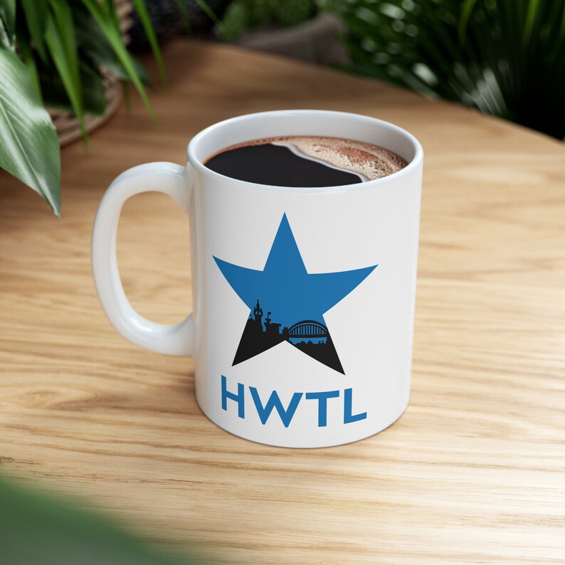 May include: White ceramic mug featuring a blue star design. Inside the star is a black silhouette of a city skyline and bridge. The letters "HWTL" are printed below the star. The mug is filled with dark coffee.