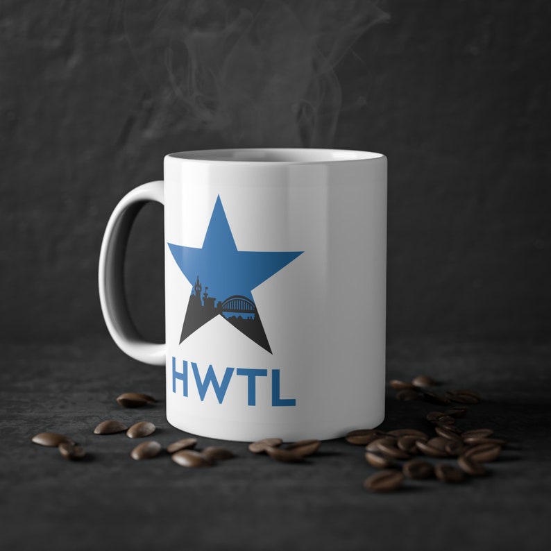May include: White ceramic mug with a blue star design, featuring a city skyline silhouette. The mug has the letters "HWTL" in blue. Coffee beans are scattered around the base of the mug, and steam rises from the top.