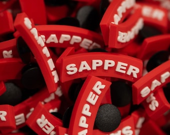 Peut inclure: Gros plan sur un tas de breloques de chaussures en caoutchouc rouge avec le mot "SAPPER" imprimé en lettres blanches.