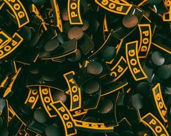 Peut inclure: Gros plan sur un tas de POGs en plastique noir avec des lettres jaunes. Les POGs sont tous en forme de rectangle incurvé aux coins arrondis. Les lettres sur les POGs disent "POG".